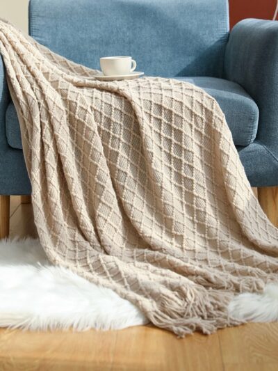 Photo d'une couverture en tricot à motifs losanges beige posé sur un fauteuil bleu avec une tasse à café et tapis en fourrure blanche sur le sol.