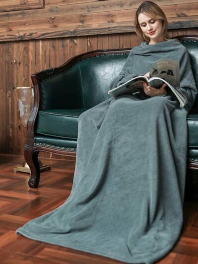 Femme lisant un journal dans un canapé vert posé sur du parquet avec une couverture grise à manches posée sur elle.