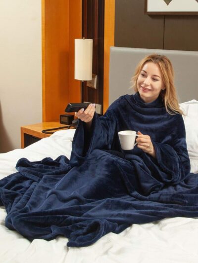 Femme assise dans un lit avec une couverture bleue sur elle, tenant une tasse blanche et une télécommande dans la main.