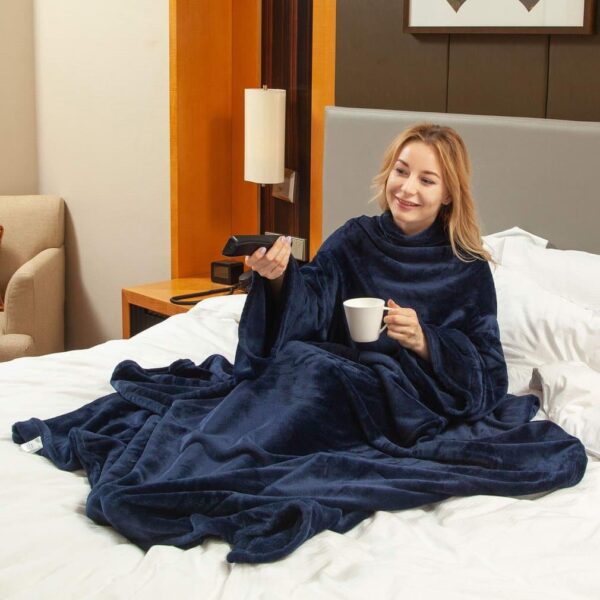 Femme assise dans un lit avec une couverture bleue sur elle, tenant une tasse blanche et une télécommande dans la main.