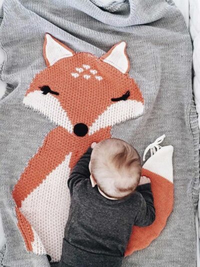 La photo est prise de haut. On voit un lit bébé dans lequel une couverture grise avec un grand motif renard est étalée. Un bébé est à quatre pattes au bout du lit.