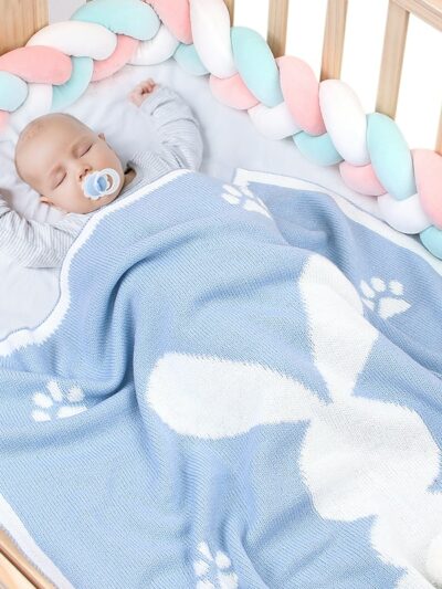On voit un bébé qui dort dans un lit à barreaux. Il est recouvert par une couverture bleue, avec un grand motif lapin. La queue du lapin est un pompon blanc qui ressort.