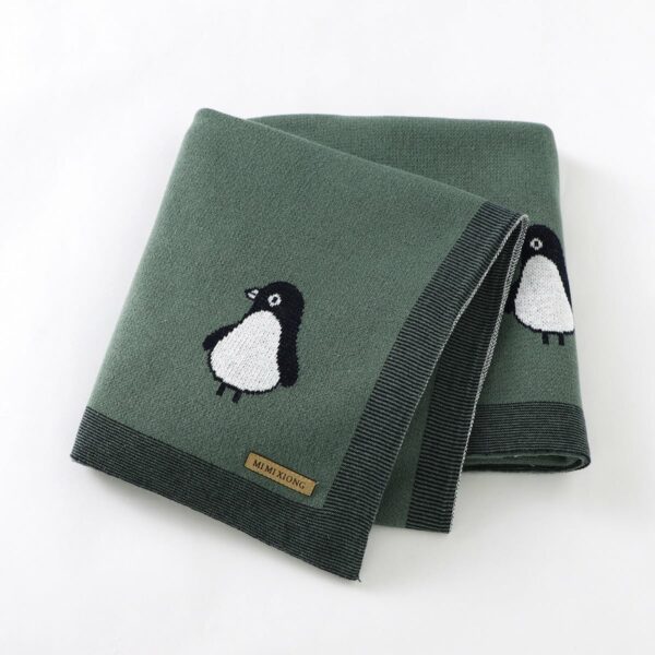 On voit une petite couverture en tricot verte ornée d'un motif pingouin très mignon. Elle est pliée en quatre et présentée sur fond blanc.