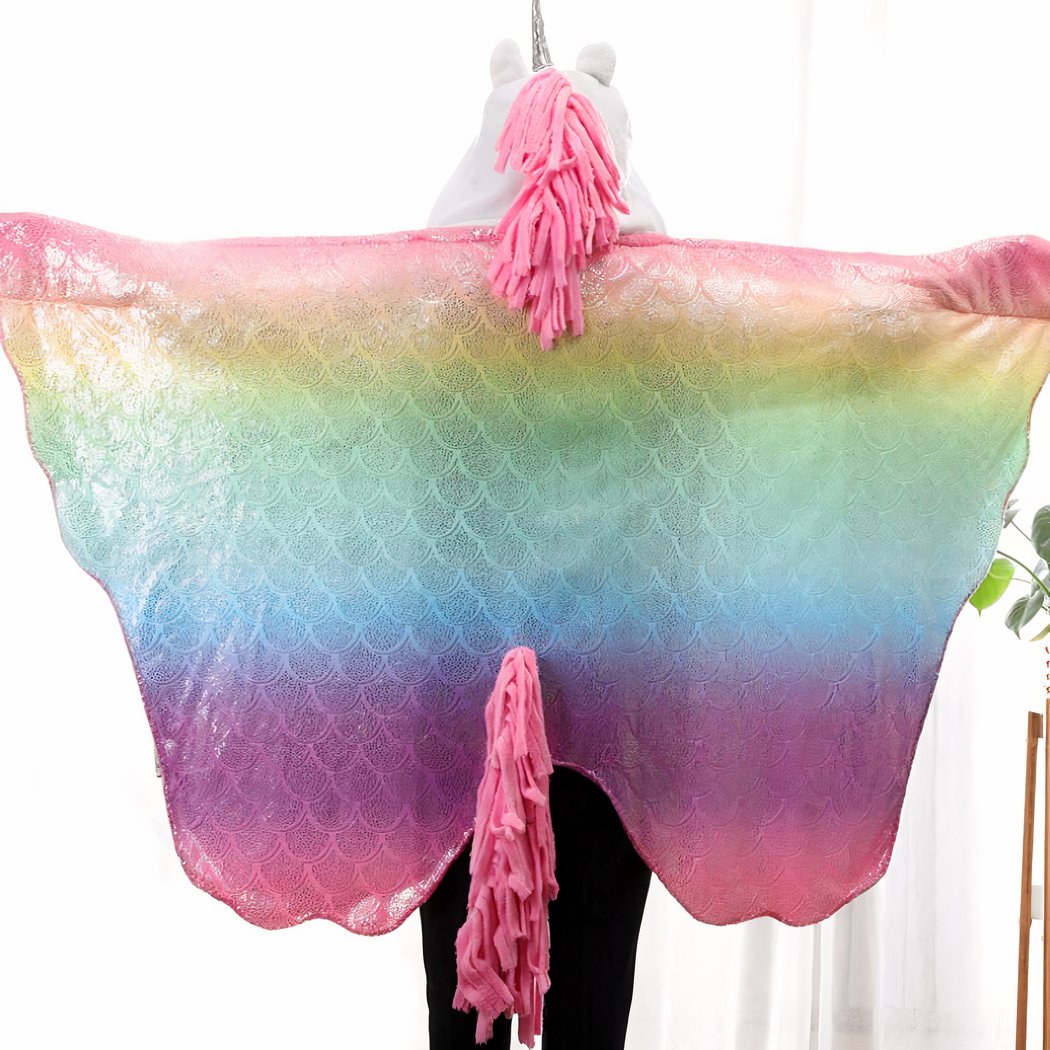 Personne de dos avec les bras écartés portant une couverture multicolore avec une crinière et queue rose ainsi qu'un corne de licorne sur la capuche