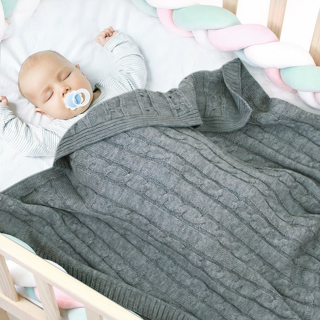L'image montre un bébé qui dort au creu de son lit à barreau. Il est recouvert d'un plaid en mailles torsadées style irlandais.