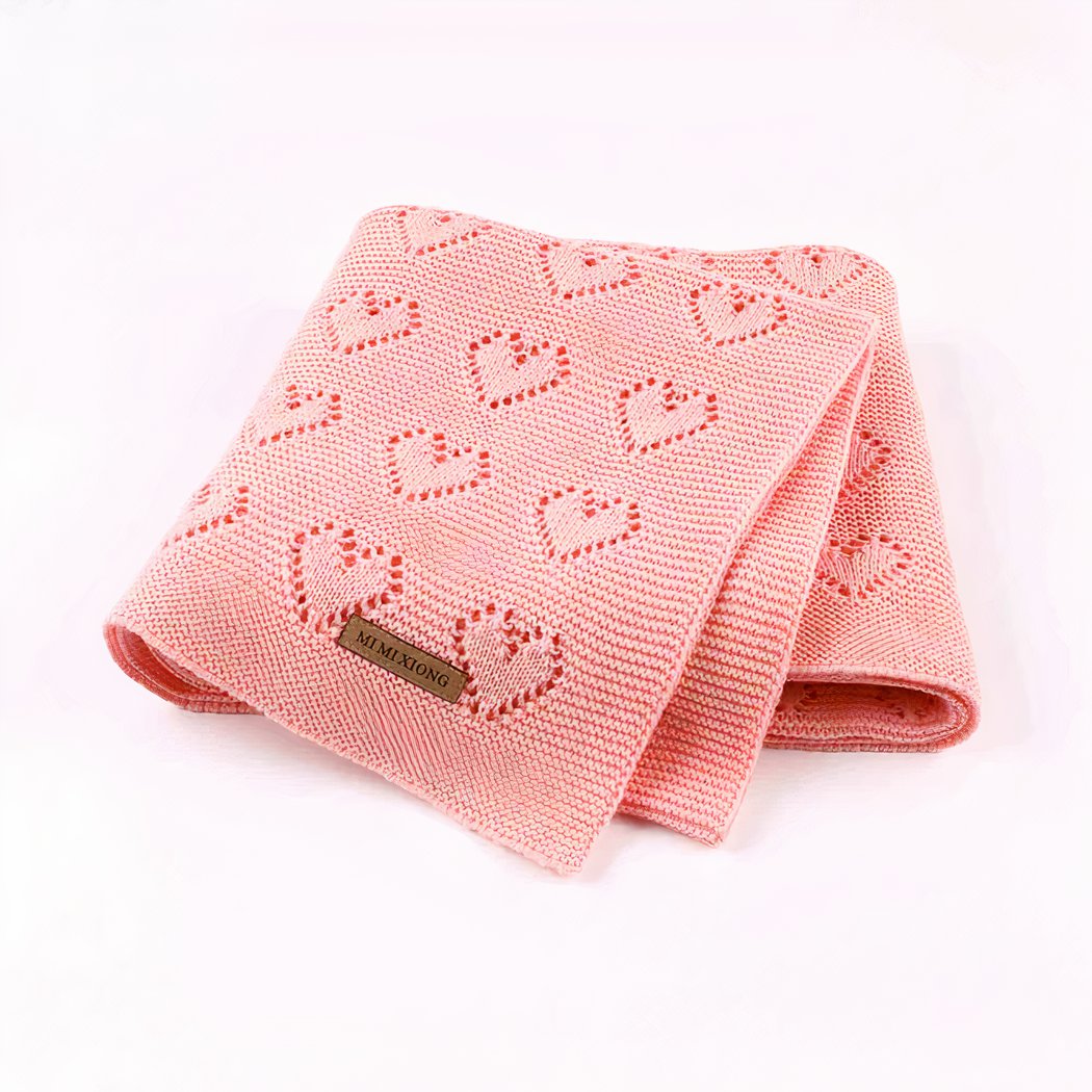 On voit une très jolie petite couverture tricotée rose à motif cœur. Elle est pliée en quatre et présentée sur fond blanc.