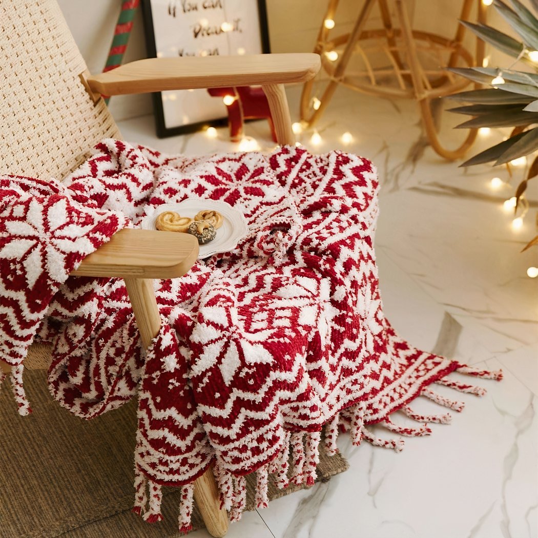Photo d'une couverture tricoté rouge et blanche avec des motifs de flocon, esprit noel. La couverture est posée sur une chaise avec une assiette de gateau.
