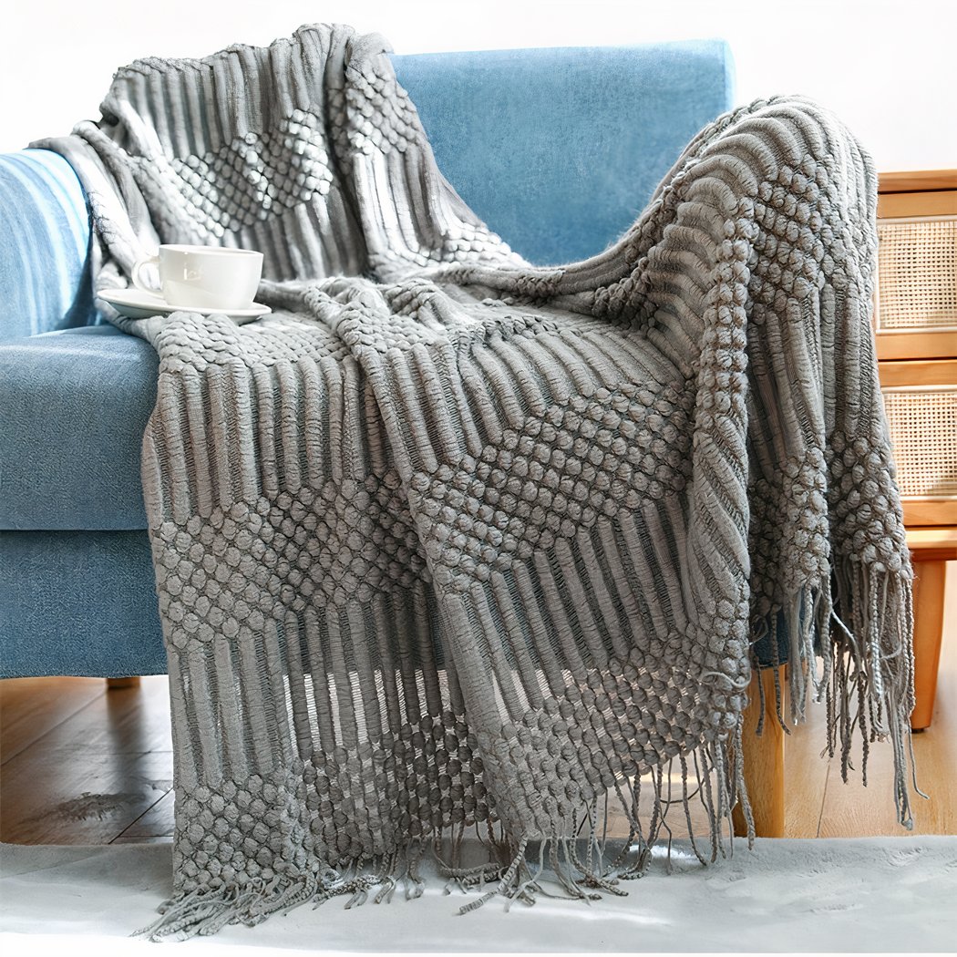 Photo d'une couverture tricotée en acrylique à motifs géormétriques texturés. La couverture en tricot grise est posé sur un fauteuil bleu avec une tasse à café dessus