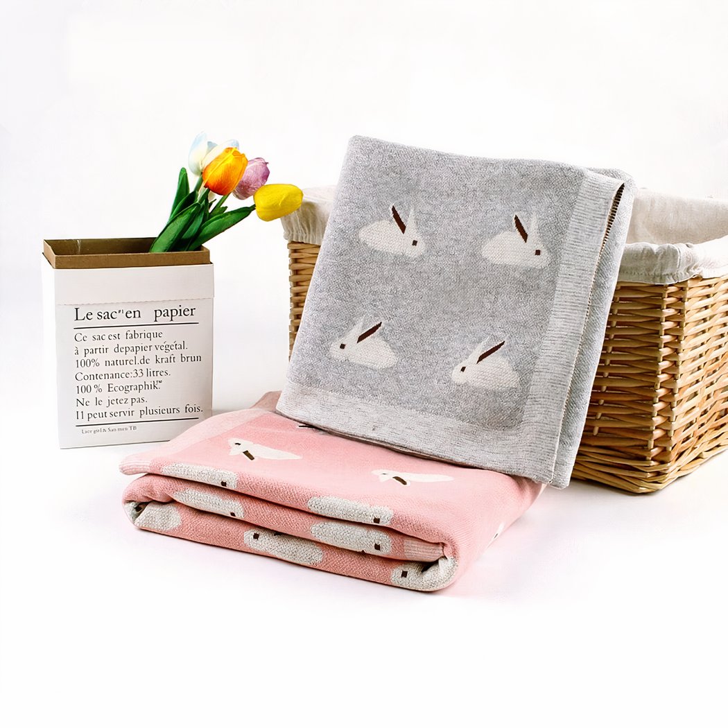 On voit deux couvertures de naissance pour bébé pliées en quatre et présentées près d'une corbeille en osier et d'un pot de fleurs rempli de tulipes. Le tout est sur fond blanc.