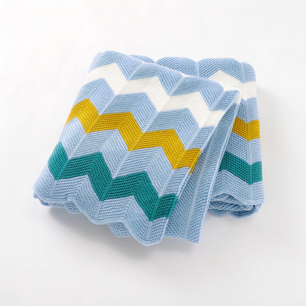 On voit une couverture pour bébé en mailles tricotées avec des motifs zigzag géographiques jaunes, blancs et bleus.
