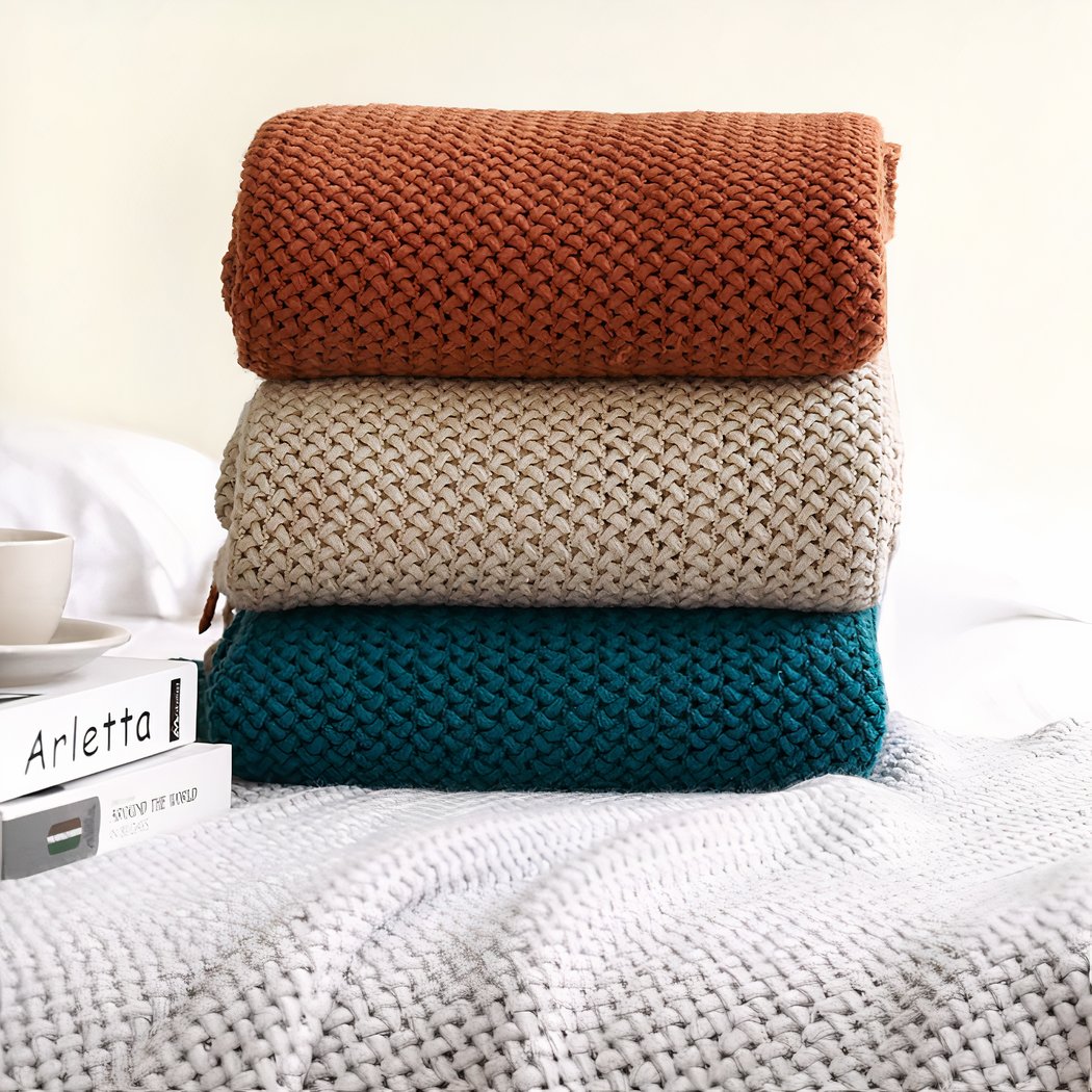 Trois couverture pliées sur un lit. Couvertures rouge, blanche et bleu unies, en mailles de laine.