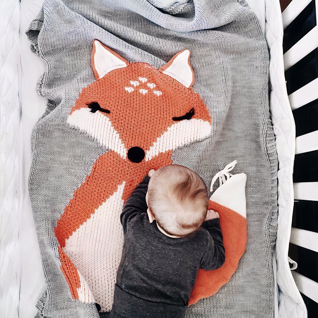 La photo est prise de haut. On voit un lit bébé dans lequel une couverture grise avec un grand motif renard est étalée. Un bébé est à quatre pattes au bout du lit.