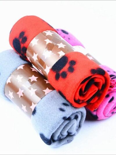 On voit trois couvertures roulées sur elles-mêmes. Elles sont rouges, roses et bleues avec des motifs pattes de chien noir. Le fond de l'image est blanc.