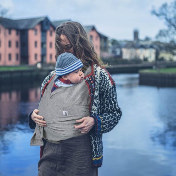Femme portant un enfant dans une couverture de portage. On voit un lac et des bâtiments au second plan.
