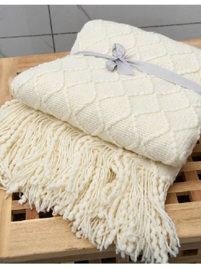 On voit une couverture en grosses mailles tricotées. Elle est beige et entourée d'un ruban gris. elle est posée sur un tabouret en bois ajouré.