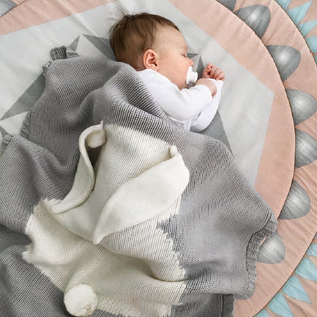 On voit un bébé brun d'environ six mois qui dort sur le flanc. Il est recouvert d'une magnifique couverture de naissance grise avec un motif lapin. Les oreilles du lapins sont en 3D et ressortent de la couverture façon doudou.