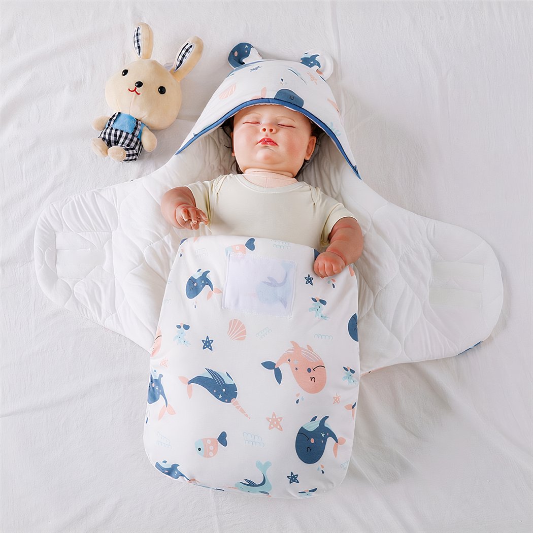 Poupée dans une couverture blanche à motifs baleines roses et bleues avec un doudou à côté de la tête du bébé.