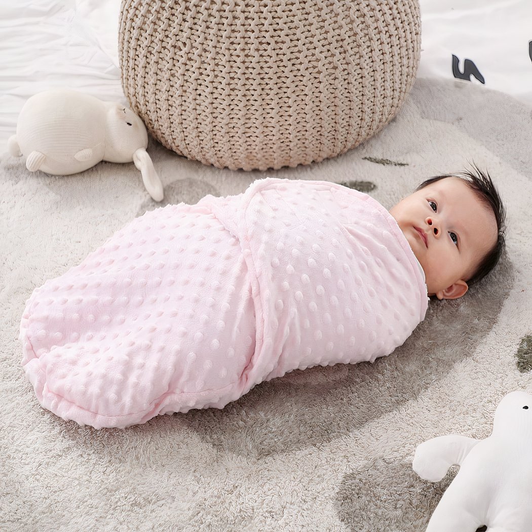 Bébé emmailloté dans une couverture rose posé sur un tapis gris avec des poufs et des doudous autour de lui
