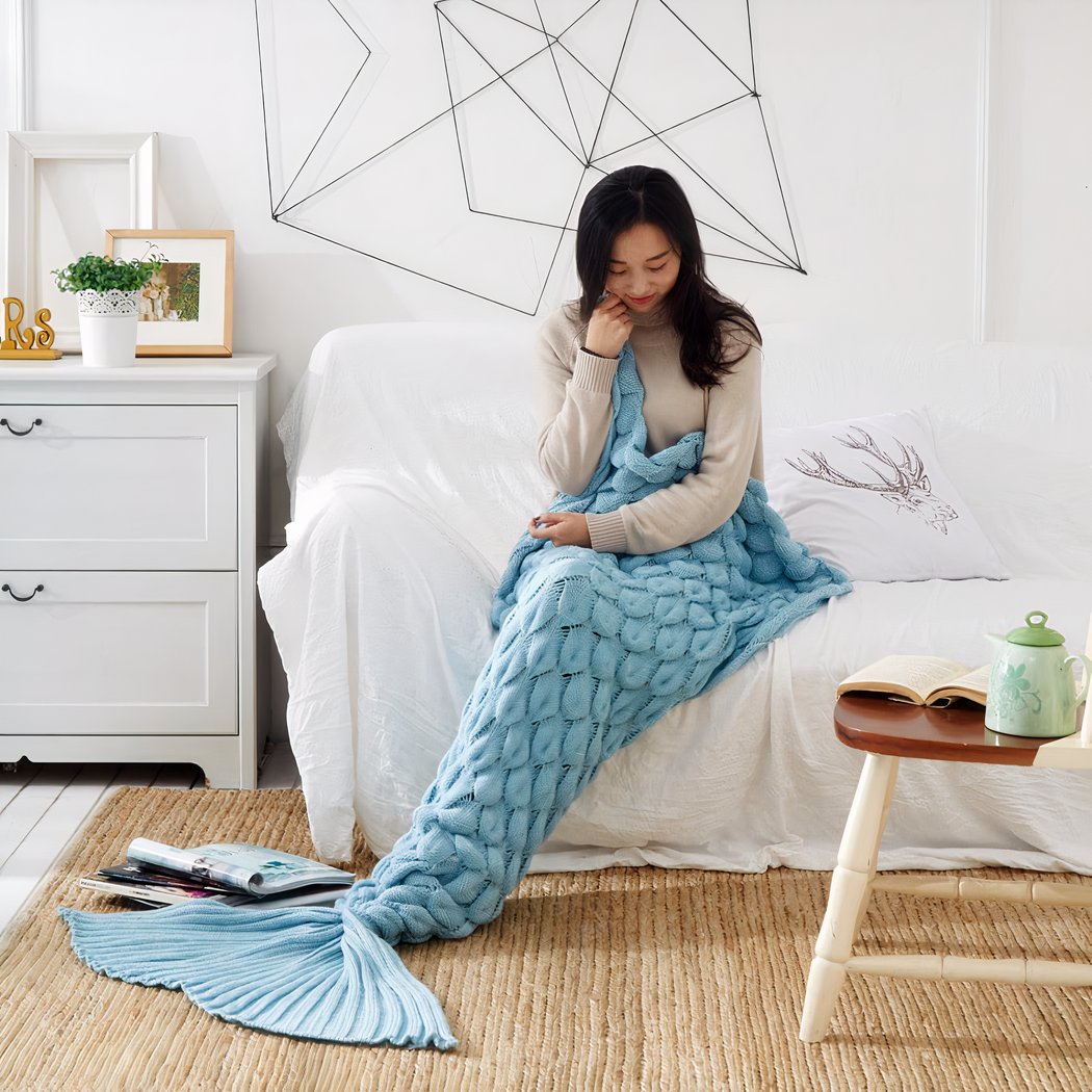 On voit une jeune femme asiatique dans une pièce blanche. Elle porte une couverture bleue avec une queue de sirène.