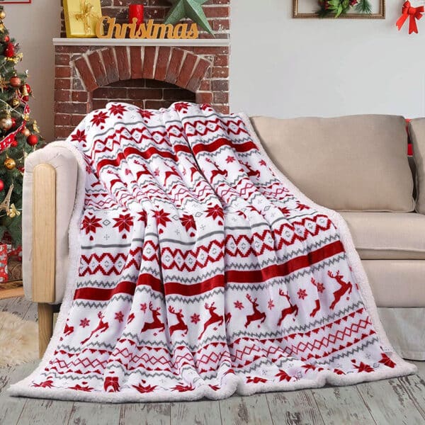 Plaid de Noël blanc et rouge comme rayé avec des motifs typiques de Noël tel que des cerfs, des flocons de neige etc. Jeter sur un canapé beige, avec une cheminée derrière et on voit un bout de sapin sur la gauche.