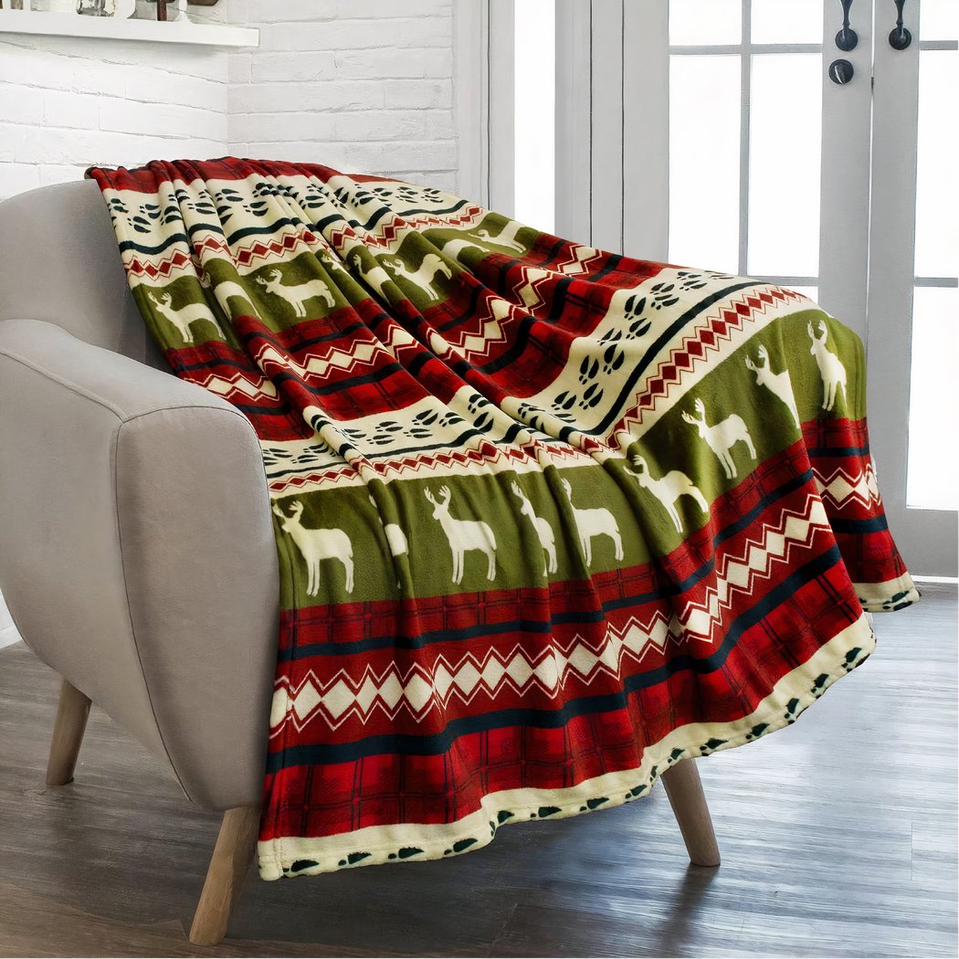 Plaid de Noël, rouge, beige et vert. Effet rayé avec des imprimés reines, losanges, lignes. Jeter sur un fauteuil gris, avec des pieds en bois. On voit du parquet au sol et dans le fond une porte-fenêtre à carreaux.